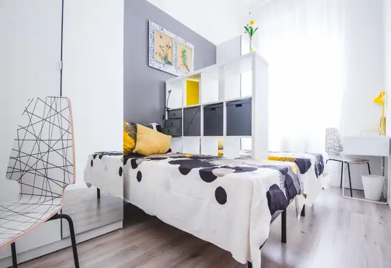 Bright, modern bedroom with two single beds separated by an open shelving unit; contemporary yellow and gray accents and natural light make the space airy and inviting.