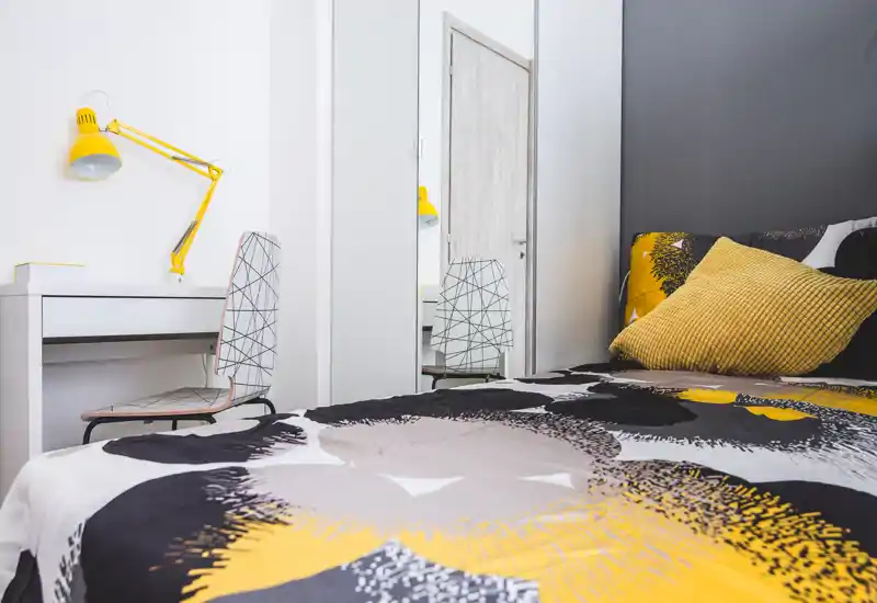 Bright, modern bedroom with a patterned duvet and warm yellow accents. A comfortable bed fills the foreground, complemented by a small desk and mirror for added functionality.
