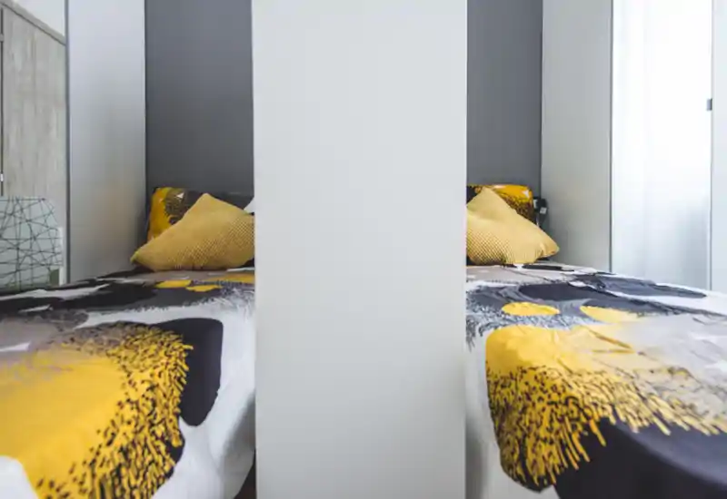Bright, modern bedroom with patterned yellow-and-gray bedding and soft cushions; natural light filters from the window creating a pleasant, tidy atmosphere.