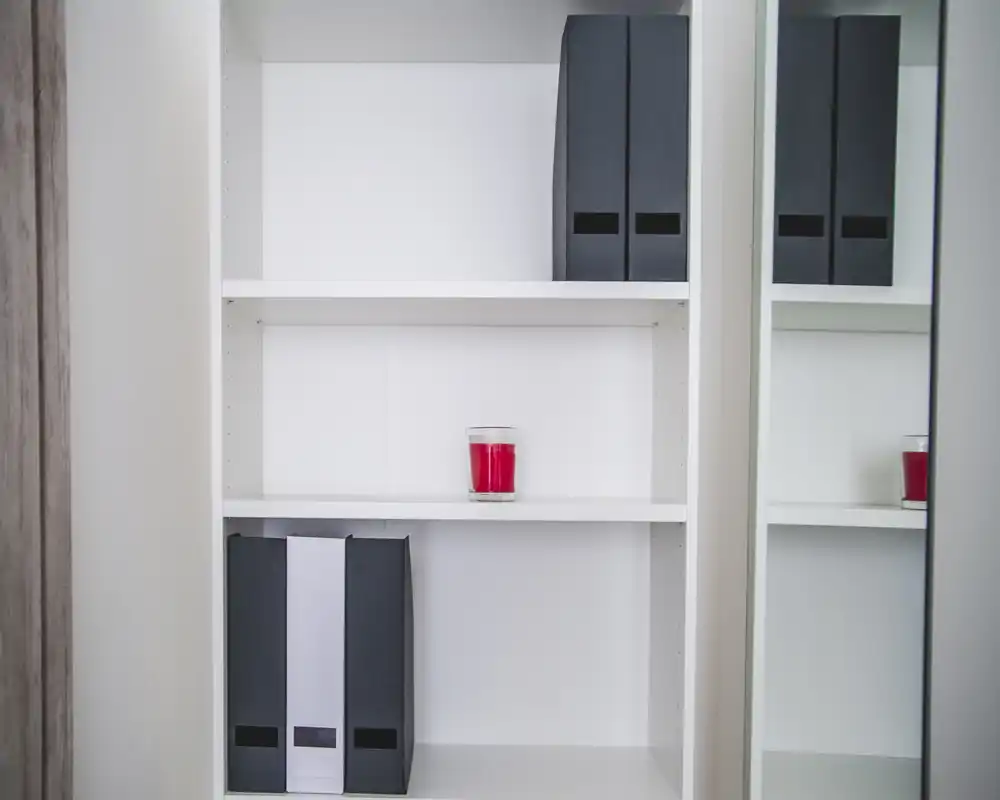 The image shows a close-up of white shelving with a few black binders and a red candle, plus a mirror reflecting part of the shelf. There are no visible features that clearly indicate a bedroom, living room, kitchen, bathroom, entrance, outdoor space, or dining area.