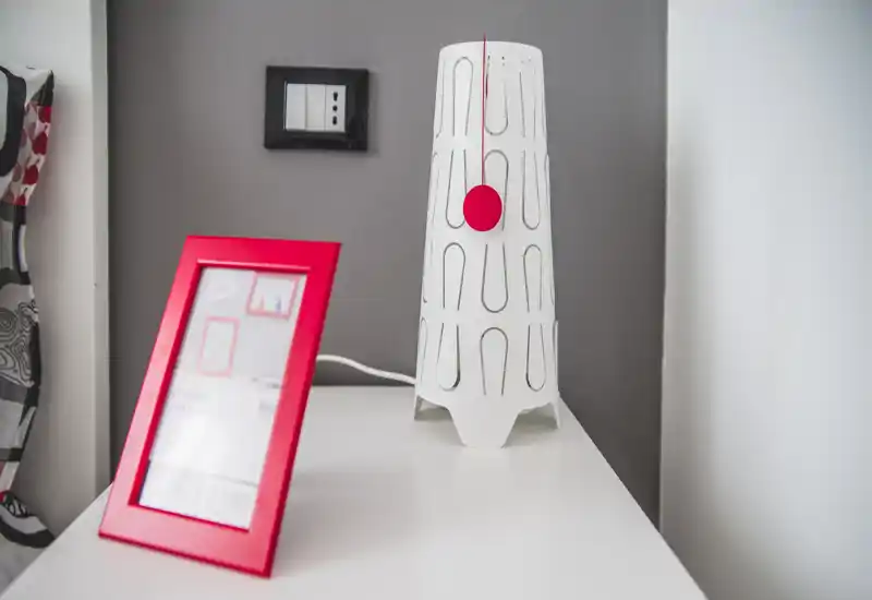 Close-up of a bedside surface with a modern white lamp and red picture frame against a neutral wall, offering a bright and tidy bedroom detail.