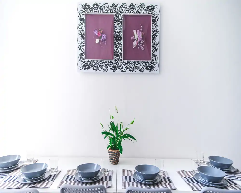 Bright, modern dining area set for multiple guests with matching grey bowls, striped placemats and a central potted plant under framed wall art.