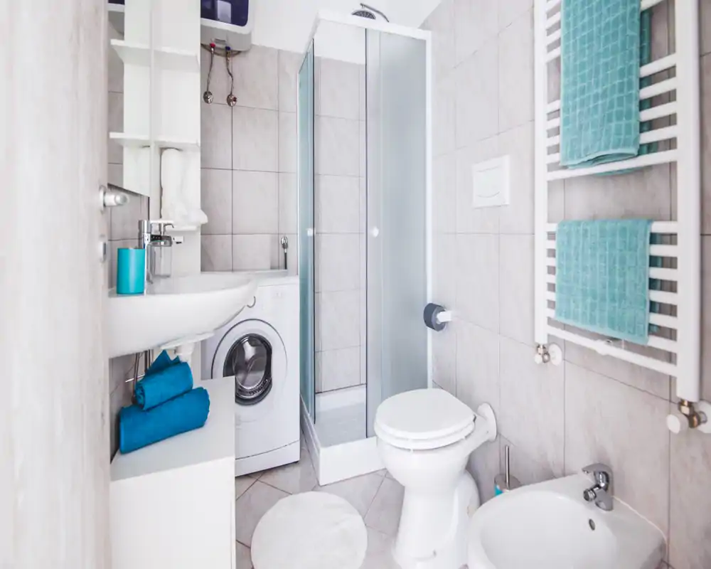 Bright, compact bathroom with a shower cabin, toilet, bidet, sink and a washing machine; light gray tiles and teal towels give a fresh, clean look.