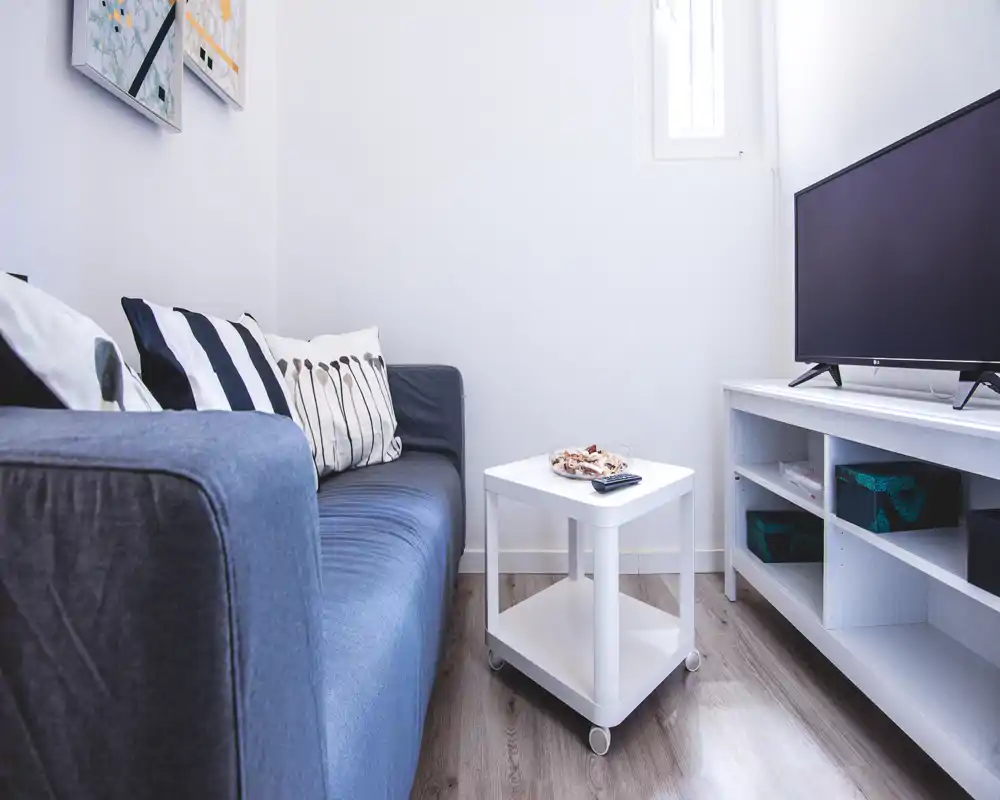 Bright, compact living area with a comfortable navy sofa, decorative cushions and a small white coffee table facing a TV unit — ideal for relaxing in a cozy, well-styled space.