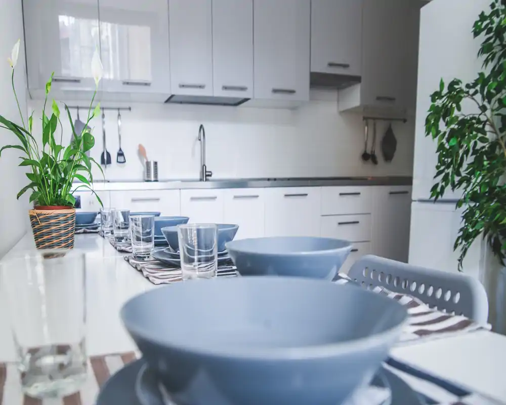 Bright modern kitchen with a long dining surface set for a meal; blue bowls, glasses and simple white cabinetry create a clean, inviting space with plants for a touch of greenery.
