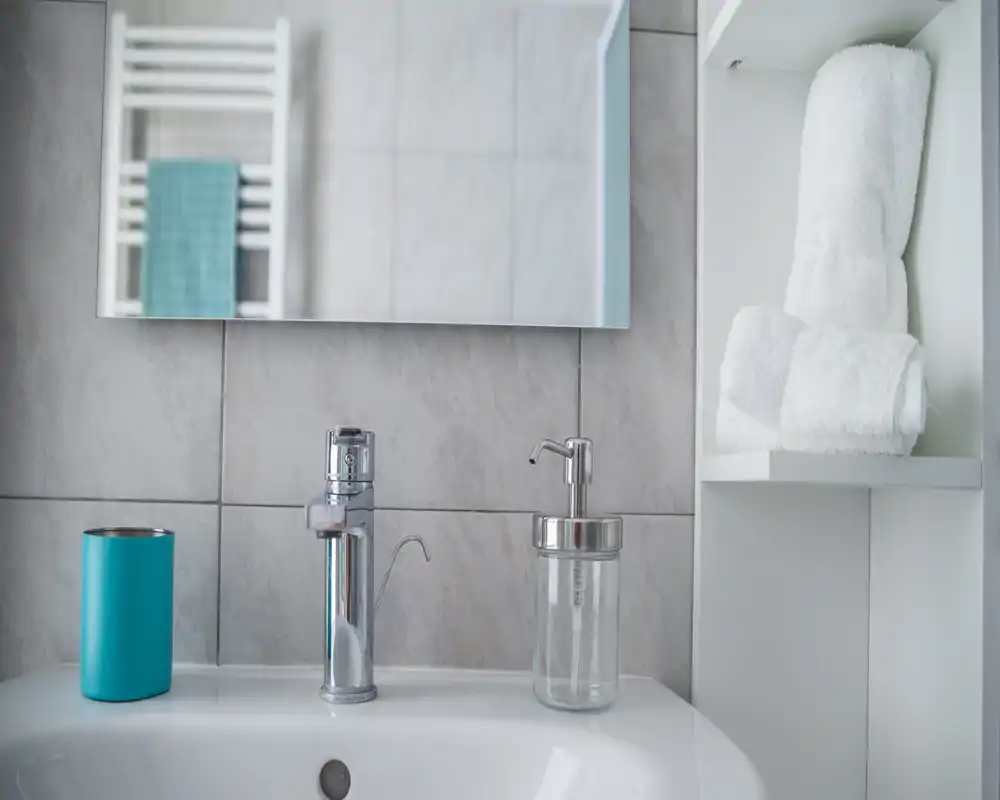 Clean bathroom sink area with a chrome faucet, soap dispenser, mirror and neatly folded white towels on a shelf.