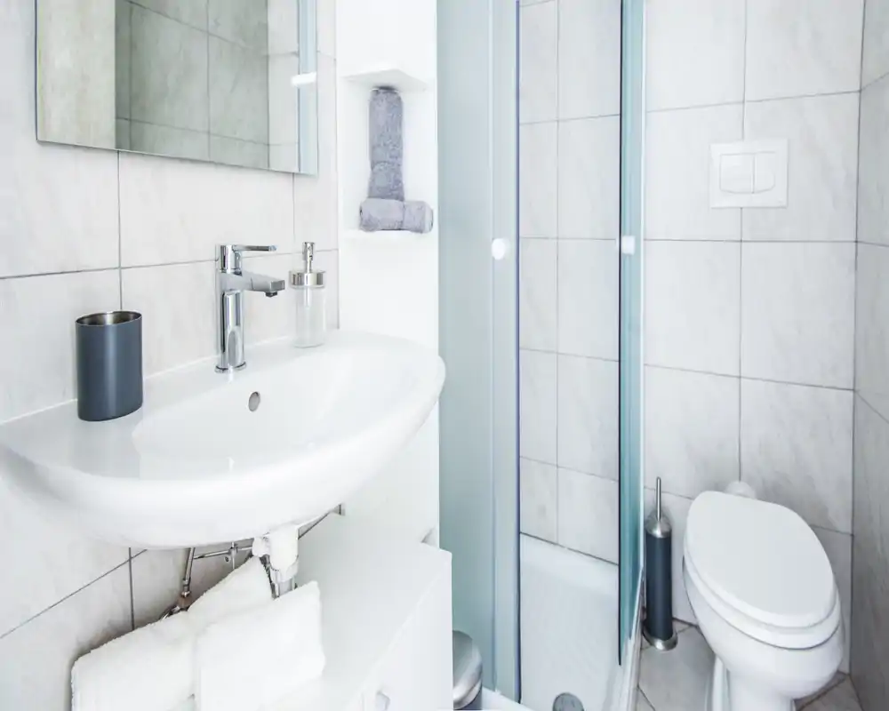 Bright, compact bathroom with a wall-mounted sink, glass shower enclosure and toilet; clean white tiles and modern fixtures.