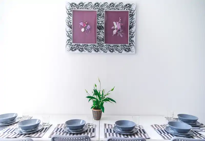 Bright, modern dining area set for multiple guests with matching grey bowls, striped placemats and a central potted plant under framed wall art.