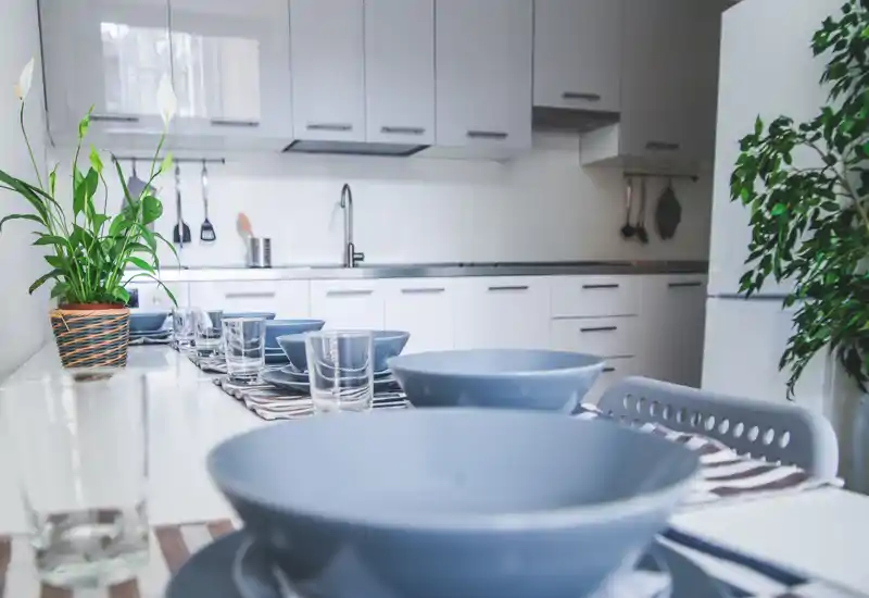 Bright modern kitchen with a long dining surface set for a meal; blue bowls, glasses and simple white cabinetry create a clean, inviting space with plants for a touch of greenery.