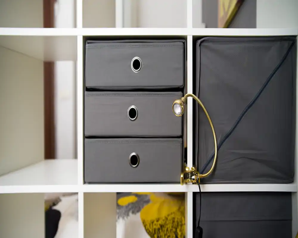 Primer plano de una estantería modular con cajas de tela grises y una pequeña lámpara ajustable; no se puede identificar claramente el tipo de habitación.
