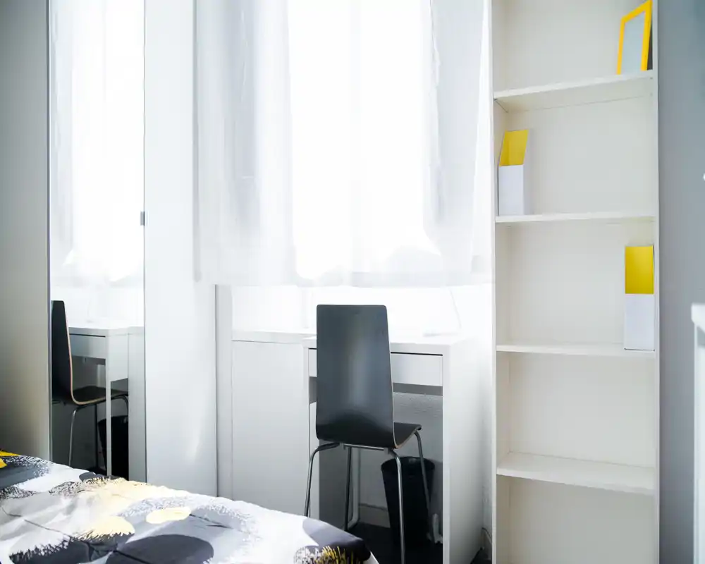 Dormitorio luminoso y minimalista con una cama con estampado en primer plano, un escritorio sencillo junto a una gran ventana y una estantería abierta. La luz natural y las superficies blancas hacen que el espacio sea fresco y acogedor, perfecto para descansar o trabajar.