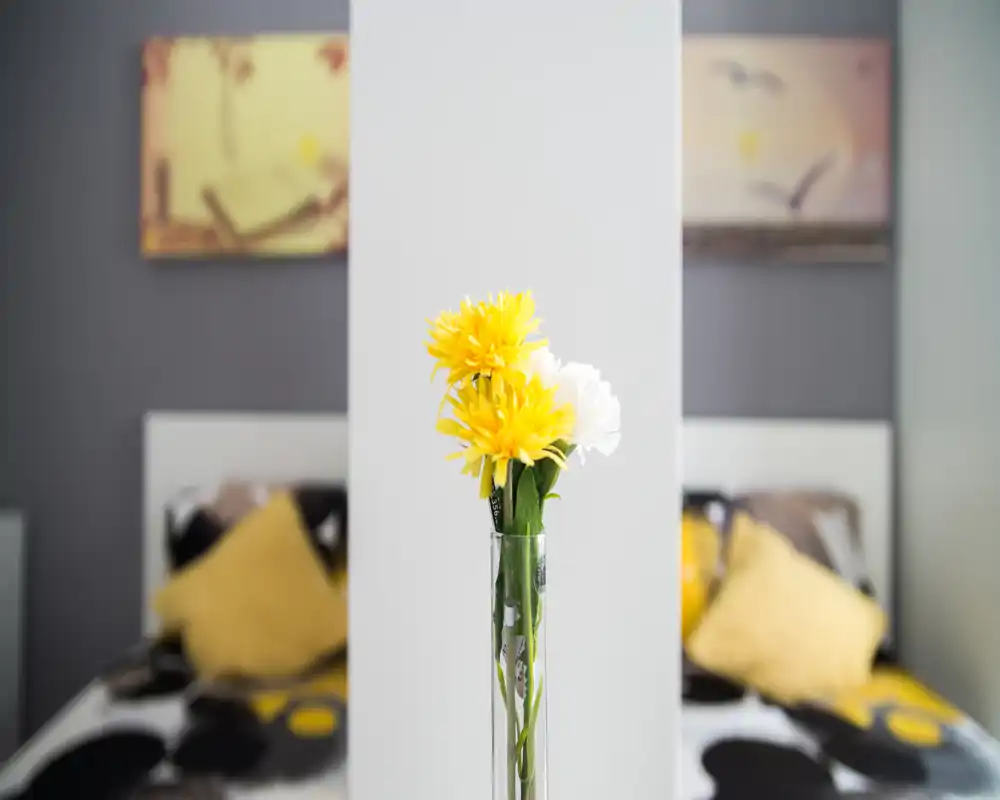 Escena de dormitorio con luz suave, un separador central y un jarrón con flores amarillas en primer plano; camas gemelas con cojines amarillos desenfocados al fondo, sensación acogedora y moderna.