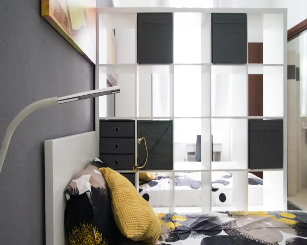 Rincón de dormitorio moderno con colcha estampada, cojines mostaza y cabecero blanco; una estantería de cubos abierta separa la cama de un área de trabajo luminosa al fondo.