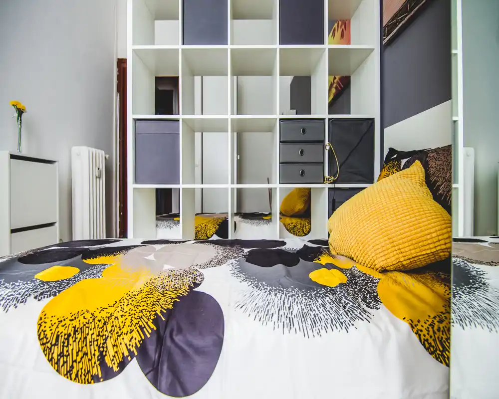Dormitorio moderno y luminoso centrado en una cama con estampado y cojines mostaza, con una estantería modular detrás; estilo limpio y contemporáneo, perfecto para anuncios.