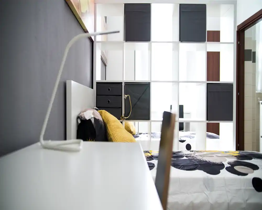 Dormitorio moderno y luminoso con ropa de cama estampada, cojines mostaza y estanterías modulares como separador; buena luz natural y aspecto contemporáneo ordenado.