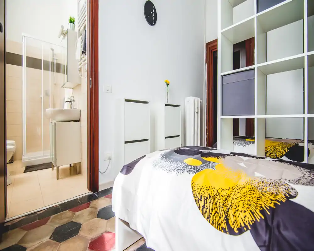 Dormitorio compacto y luminoso con edredón estampado, estantería espejo para almacenamiento y baño en suite visible por la puerta.