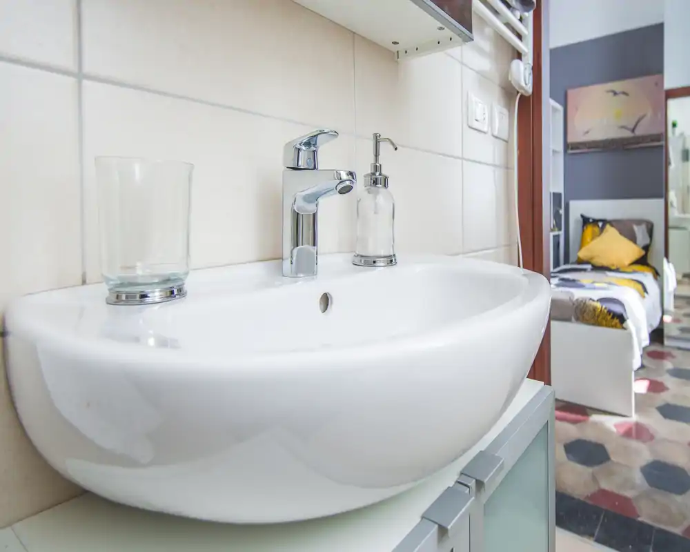 Primer plano de un lavabo de baño limpio con grifo moderno y dosificador de jabón; se aprecia un dormitorio al fondo a través de la puerta.