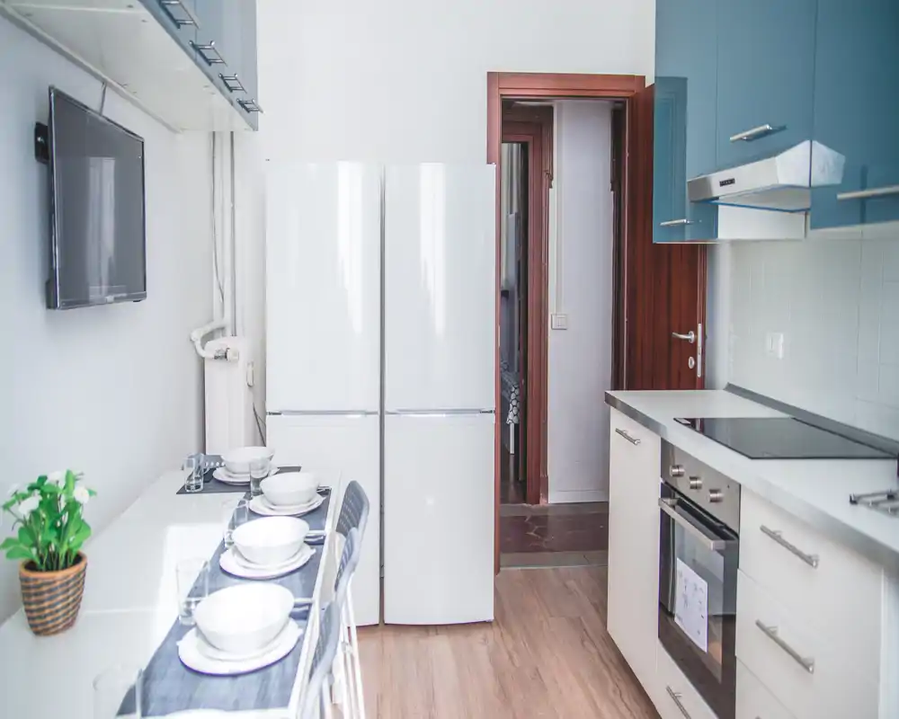 Cocina moderna equipada con horno empotrado, placa de inducción y frigorífico de dos puertas; pequeña mesa para cuatro y ambiente luminoso.