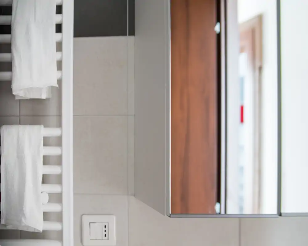 Primer plano de un rincón de baño moderno con toallero radiador y toallas blancas, pared alicatada y armario con espejo que refleja una puerta de madera.