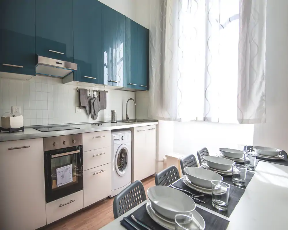 Cocina moderna y luminosa con armarios superiores brillantes, horno integrado y lavadora, y un área de comedor puesta junto a una gran ventana.