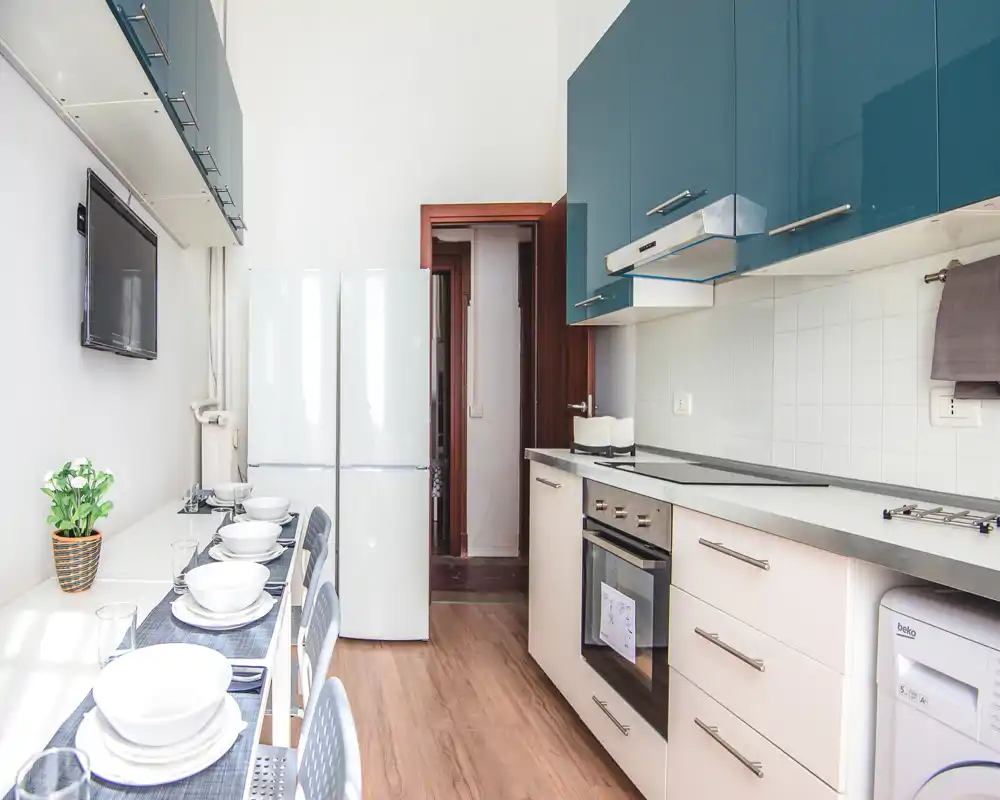 Cocina moderna y luminosa con armarios altos verde azulado, horno empotrado, lavavajillas y un pequeño mostrador-comedor para cuatro. Líneas limpias, suelo de madera y luz natural crean un espacio funcional y acogedor, perfecto para alquileres temporales.