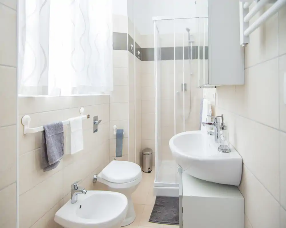 Baño compacto y luminoso con lavabo mural, bidé, inodoro y ducha en esquina; azulejos neutros y luz natural desde la ventana con cortina que aportan una sensación limpia y aireada.