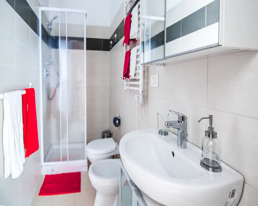 Baño moderno y compacto con lavabo, inodoro, bidé y ducha cerrada. Baldosas neutras y acentos rojos que ofrecen una imagen limpia y cuidada, perfecta para anuncios de alquiler.