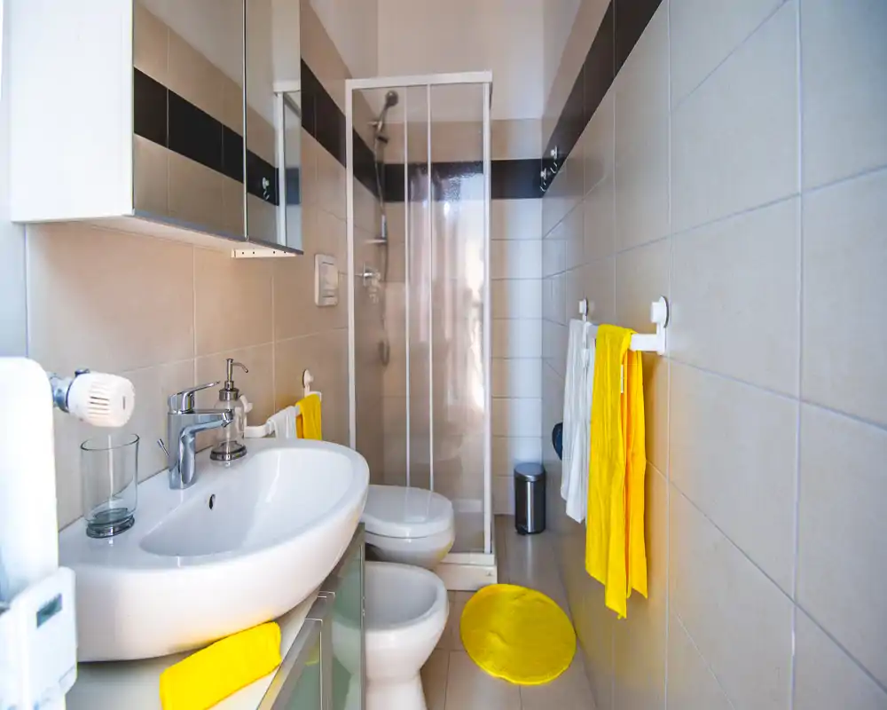 Baño luminoso y cuidado con lavabo, bidé, inodoro y ducha en esquina; toallas amarillas y alfombrilla a juego aportan un toque alegre.