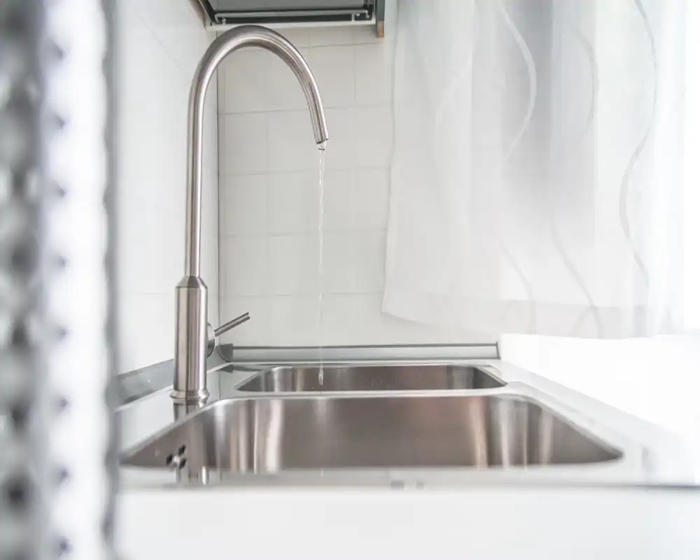Fregadero de cocina luminoso y minimalista con grifo moderno de acero inoxidable y doble seno. La luz natural y una cortina translúcida crean una atmósfera limpia y aireada, ideal para destacar detalles.