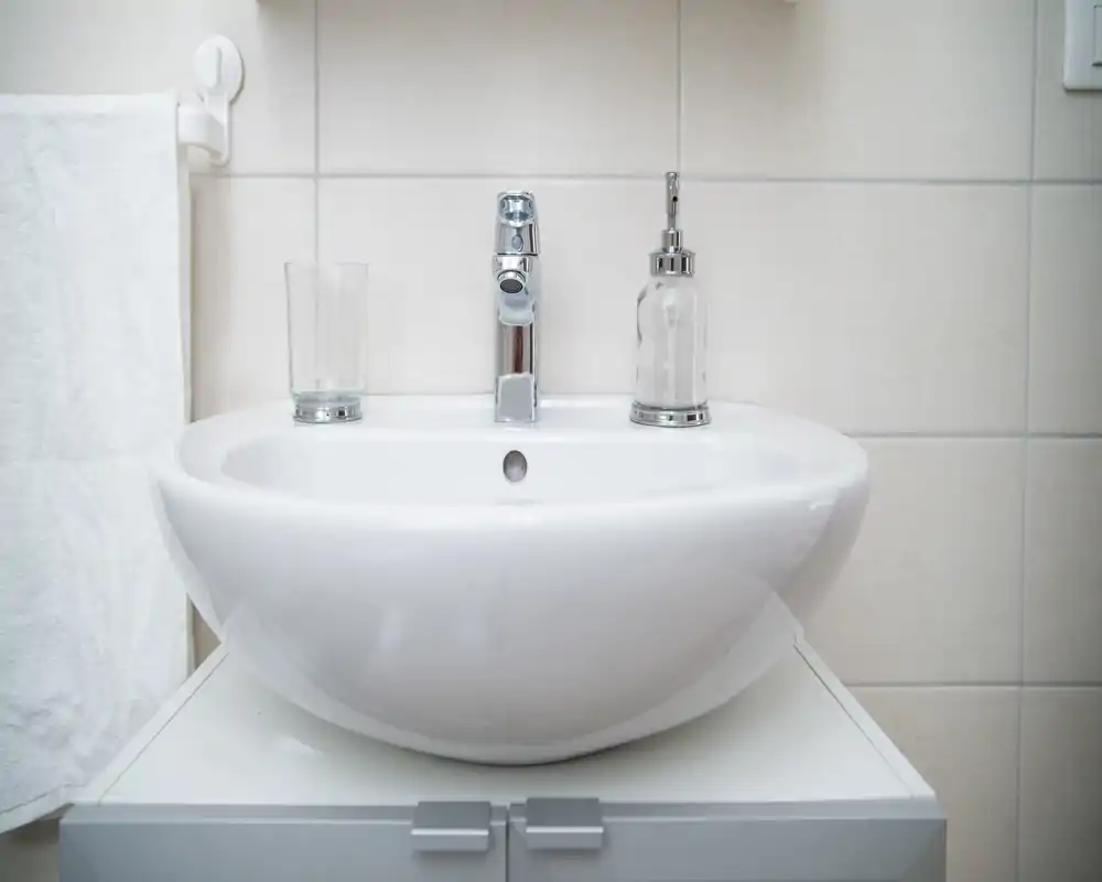 Lavabo de baño moderno y limpio con grifo cromado, dosificador de jabón y vaso, sobre pared de azulejos — luminoso y bien cuidado.