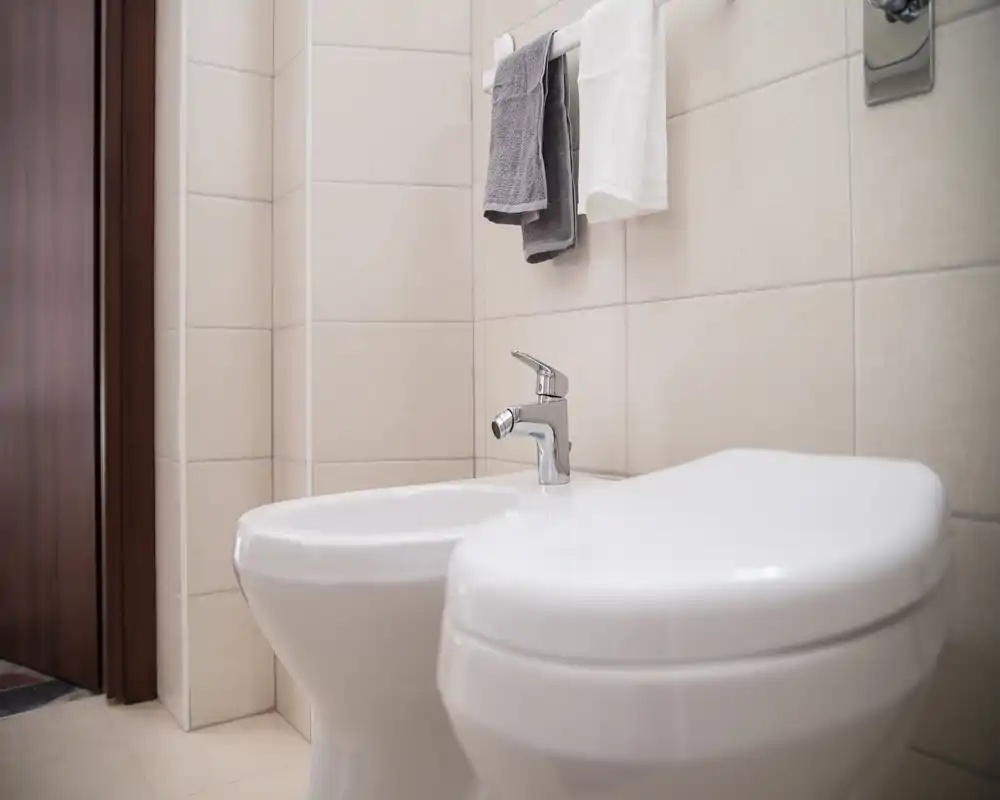 Baño moderno y compacto con inodoro y bidé, paredes alicatadas y toallero; limpio, minimalista y bien iluminado.