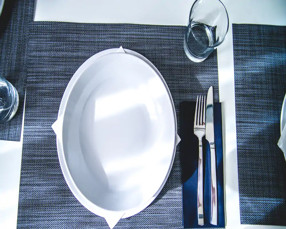 Vista cenital de un puesto en la mesa bien dispuesto con un plato oval blanco, cubiertos sobre una servilleta oscura y un vaso sobre un salvamantel tejido.
