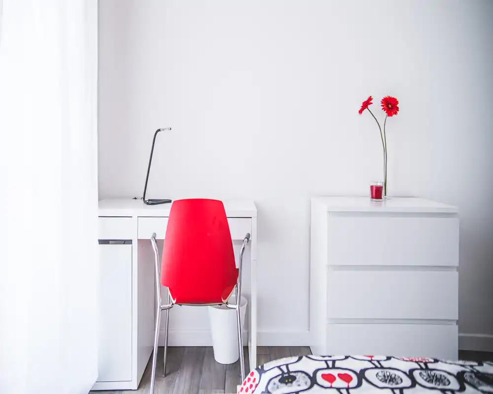 Rincón de dormitorio luminoso y minimalista con escritorio blanco, silla roja y cómoda a juego; una colcha estampada asoma en la imagen y dos flores rojas dan color.