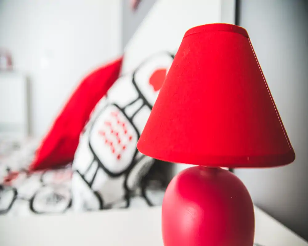 Plano detalle de la mesita de noche con una lámpara roja vibrante y la cama desenfocada con cojines estampados al fondo, que aporta una sensación acogedora y moderna.