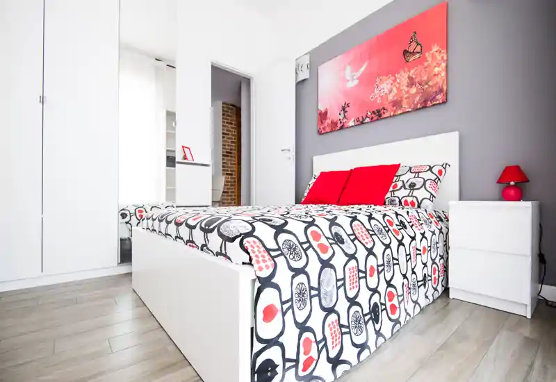 Dormitorio moderno y luminoso con cama doble estampada, cojines rojos y cuadro a juego; mobiliario blanco y luz natural que crean un ambiente acogedor.