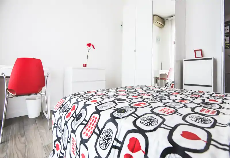 Dormitorio moderno y luminoso con ropa de cama estampada, armario blanco y silla roja como acento; estilo minimalista y luz natural.
