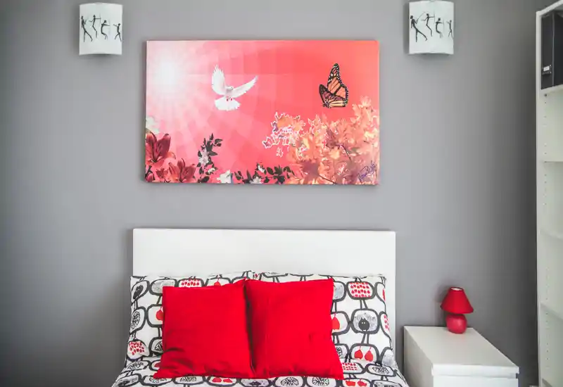 Dormitorio luminoso y con estilo, con cabecero blanco, ropa de cama estampada y cojines rojos llamativos; cuadro y detalles coordinados crean un ambiente moderno y acogedor.