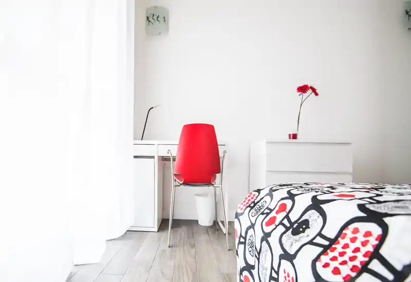 Rincón de dormitorio luminoso y minimalista con colcha estampada, escritorio blanco y silla roja llamativa; una flor roja añade un toque decorativo y la luz natural inunda el espacio.