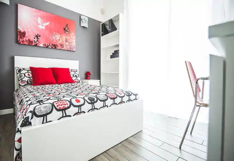 Dormitorio moderno y luminoso con colcha estampada, cojines rojos y una cama blanca minimalista. Gran ventana con cortinas translúcidas que llena la habitación de luz natural y un cuadro que realza la pared gris.