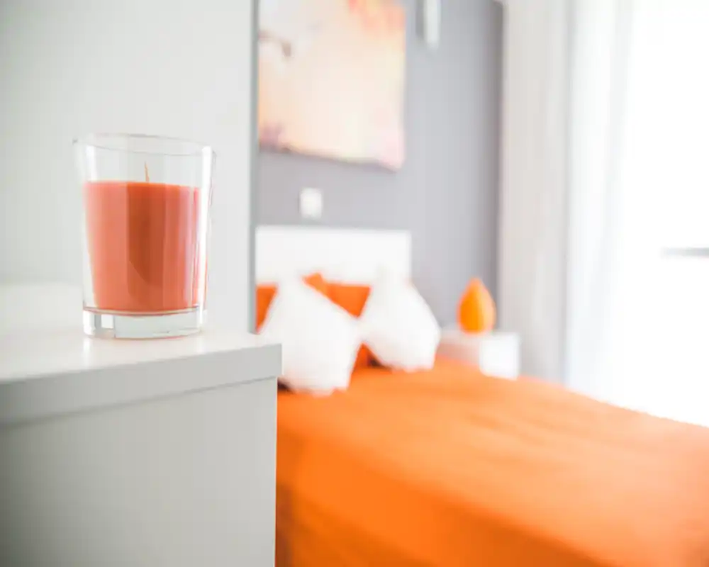 Camera da letto luminosa e moderna con copriletto arancione, testiera bianca e cuscini decorativi; dettagli caldi come una candela coordinata rendono l'ambiente accogliente.