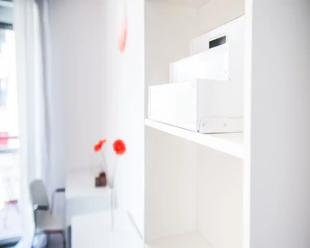 Zona giorno luminosa e minimal con scaffalatura bianca, una scrivania e fiori decorativi; composizione ariosa e leggermente morbida con luce naturale dalla finestra.