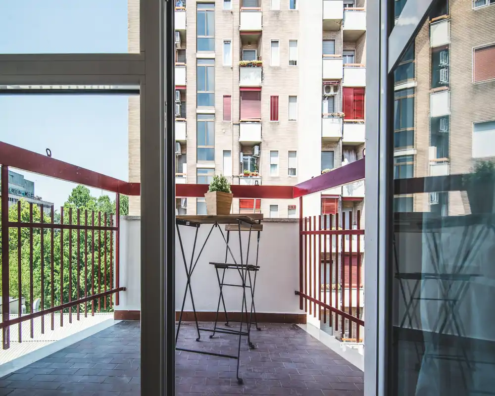 Balcone urbano soleggiato con piccolo tavolo e sedie pieghevoli, ringhiera rossa e vista sulla città — perfetto per caffè mattutino.