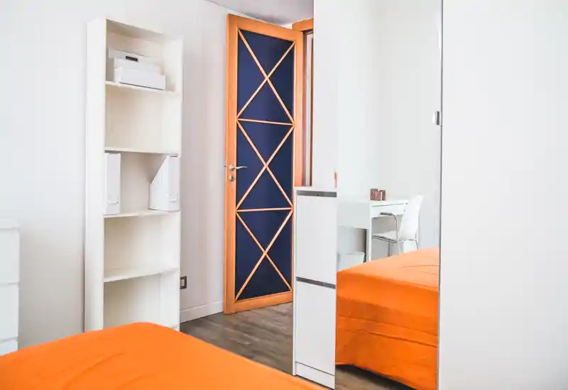 Angolo di camera luminoso e moderno con copriletto arancione, scaffalatura bianca e guardaroba con specchio che riflette il letto e una piccola postazione.