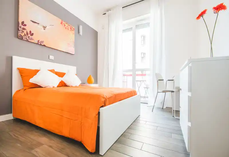 Camera da letto luminosa e moderna con letto dalla struttura bianca e biancheria arancione vivace, cuscini morbidi e luce naturale da una grande finestra/portafinestra — pavimento in legno chiaro e arredo minimal.