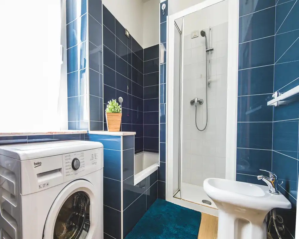 Baño moderno y luminoso con azulejos azules, ducha, bidé y lavadora; espacio compacto y bien iluminado con alfombra y una pequeña planta.