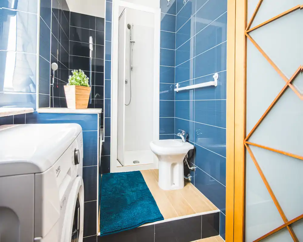 Bagno moderno e luminoso con pareti piastrellate blu, doccia ad angolo, bidet e lavatrice; pavimento effetto legno e tappetino turchese per un tocco caldo.