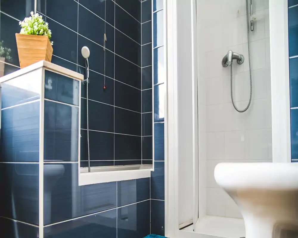 Baño moderno y compacto con azulejos azul oscuro, ducha con mampara de cristal, cabezales de ducha de mano y un inodoro blanco en primer plano.