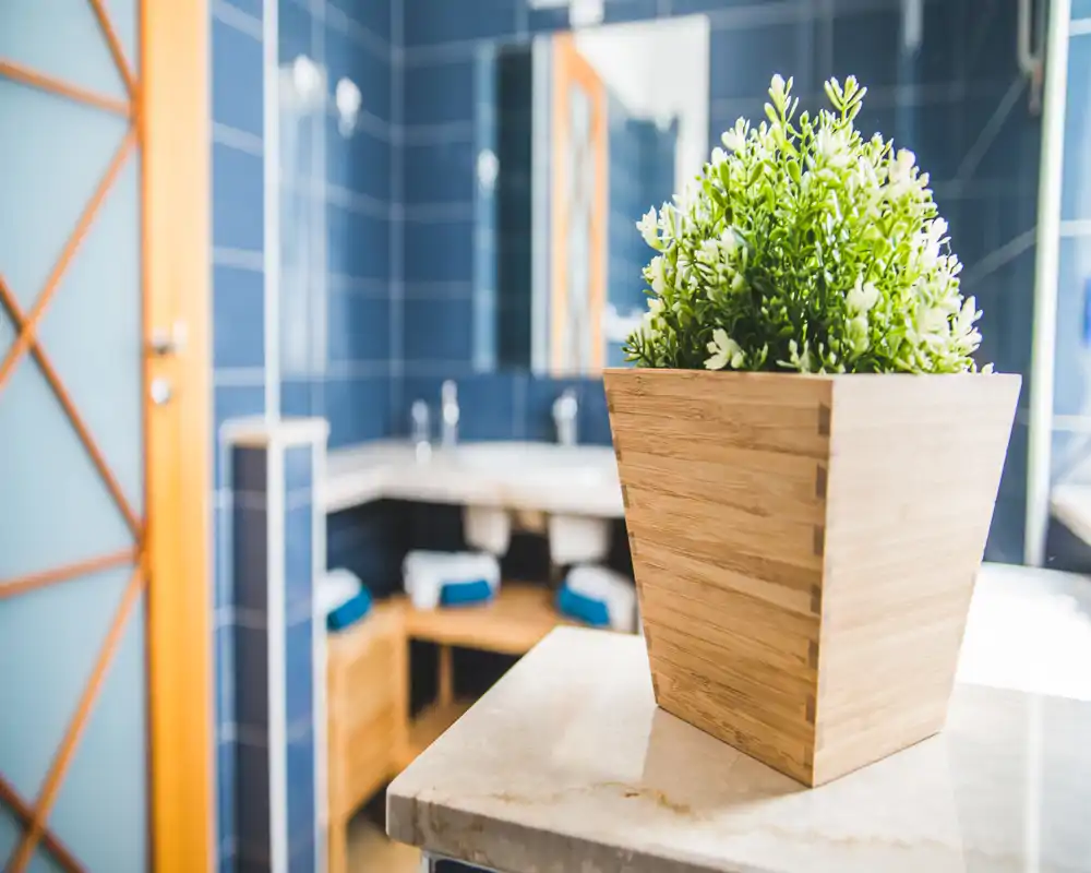 Detalle de un baño luminoso y con estilo, con una maceta de madera en primer plano y el área del lavabo con azulejos visible al fondo.