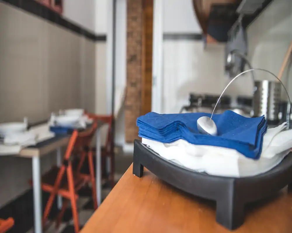 Primer plano de una encimera de cocina con servilletas apiladas y utensilios; al fondo, desenfocado, se ve un pequeño comedor con sillas plegables.