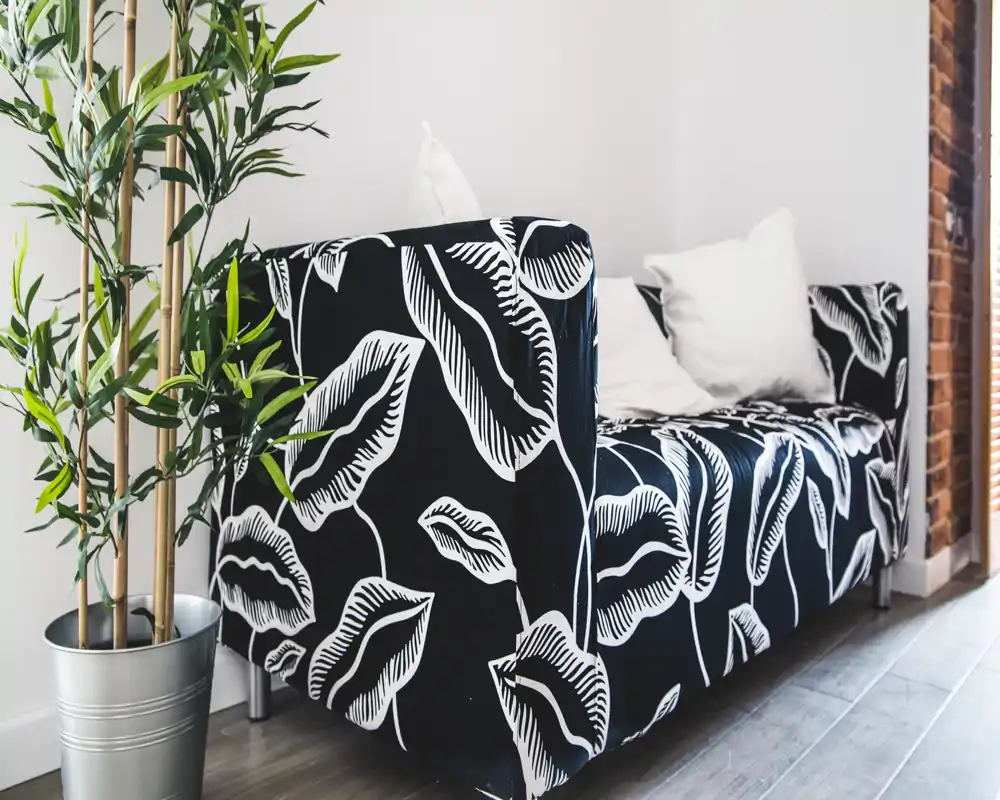 Zona de estar con estilo que muestra un sofá estampado en blanco y negro, cojines y una planta en maceta, iluminada por luz natural — un rincón acogedor y con diseño.